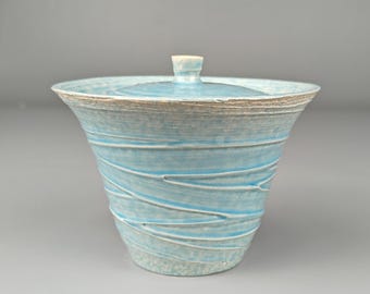 May include: A light blue ceramic jar with a lid. The jar has a wide, flared shape with a textured, swirling pattern. The lid has a small, rounded handle. The rim of the jar and lid have a contrasting, light brown color.
