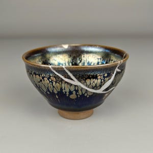 May include: A ceramic bowl with a dark blue and gold iridescent glaze. The bowl has a small, round base and a white, vein-like repair. The interior has a speckled pattern. The rim is a dark brown color.