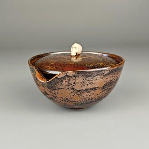 Könnte beinhalten: Eine braune Keramikschale mit Deckel und einem kleinen, schädelartigen Griff. Die Schale hat eine strukturierte, gesprenkelte Oberfläche mit dunkelbraunen und schwarzen Streifen. Der Deckel passt gut, und die Schale hat einen Ausguss.