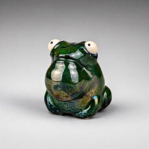 Peut inclure: Figurine de grenouille en céramique avec un vernis vert foncé brillant. La grenouille a de grands yeux blancs et un corps arrondi. L'objet est décoratif.