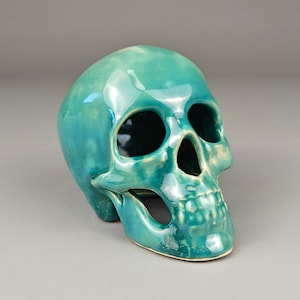 May include: A turquoise ceramic skull sculpture with a glossy finish. The skull has large eye sockets, a prominent nose cavity, and a detailed jawline. The sculpture is a decorative item, likely for Halloween or gothic decor.