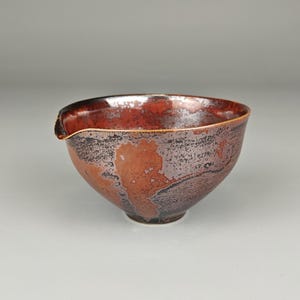 Pode incluir: Uma tigela de cerâmica artesanal com uma superfície texturizada única. A tigela apresenta uma rica paleta de cores vermelha, castanha e preta, com um pequeno bico na borda. O design é orgânico e artístico, adequado para servir ou expor.