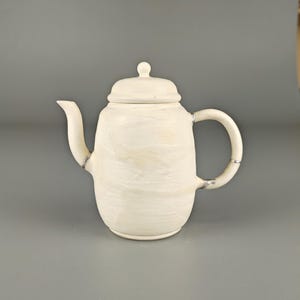 May include: Cream-colored ceramic teapot with a lid and curved handle. The teapot has a spout and a small knob on the lid. The surface has a textured, marbled appearance. A classic kitchenware item.