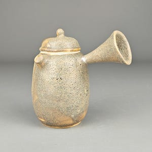May include: A ceramic teapot with a speckled, textured surface in shades of gray, brown, and beige. The teapot has a lid with a small knob, a spout, and a distinctive, flared handle. The design is rustic and artisanal.