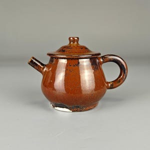 May include: A small, brown ceramic teapot with a lid and a curved handle. The teapot has a glossy finish and a spout for pouring. The design is simple and classic, suitable for serving tea. The teapot is approximately 5 inches in height.