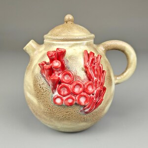 May include: A small, round teapot with a lid and a handle. The teapot is decorated with red coral-like shapes.