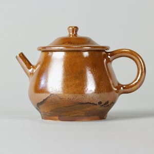 Può includere: Teiera in ceramica marrone con coperchio, beccuccio e manico. La teiera ha un corpo arrotondato e una finitura lucida. Il colore è un marrone caldo e terroso con accenti più scuri vicino alla base. Il manico è un semplice anello.
