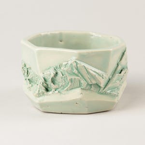 May include: A pale mint green ceramic bowl with a faceted, geometric exterior. The surface features a raised, textured relief design depicting a mountain range landscape wrapping around the sides of the vessel, finished with a glossy glaze.