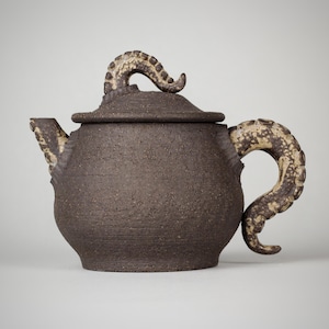 Könnte beinhalten: Eine dunkelbraune Keramik-Teekanne mit Deckel und Ausguss. Der Griff und die Oberseite des Deckels haben ein strukturiertes, tentakelartiges Design in einer helleren Brauntönung. Die Teekanne hat ein rustikales, handgemachtes Aussehen.