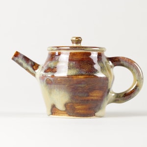 Peut inclure: Une théière en céramique faite à la main avec un couvercle. La théière présente un glaçage marron et beige, un corps arrondi, une anse incurvée et un bec verseur. La théière est conçue pour infuser du thé.