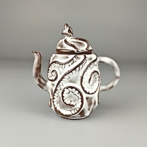 Könnte beinhalten: Eine Keramik-Teekanne mit weißem Grund und braunen Akzenten. Die Teekanne hat einen Deckel und einen Griff mit einem dekorativen Wirbelmuster. Der Ausguss ist gebogen, und das Gesamtdesign ist künstlerisch und einzigartig.