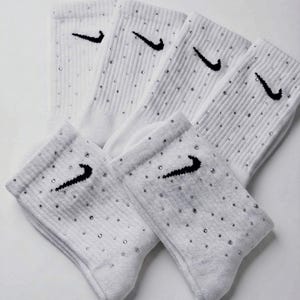 Könnte beinhalten: Weiße Sportsocken mit schwarzem Nike Swoosh-Logo und kleinen, funkelnden Verzierungen. Die Socken sind gerippt und haben eine klassische Crew-Länge. Ein stilvolles und sportliches Accessoire.