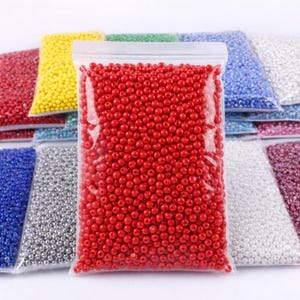 Op de afbeelding: Een doorzichtige plastic zak gevuld met kleine, ronde, felrode kralen. Andere zakken met kralen in verschillende kleuren, waaronder geel, blauw, zilver en wit, zijn zichtbaar op de achtergrond. Deze kralen zijn waarschijnlijk voor knutselen of het maken van sieraden.