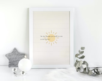 Puede incluir: Una impresión enmarcada blanca con un gráfico de sol amarillo y la cita "Sé el cambio que quieres ver en el mundo." La impresión está sobre un fondo de madera blanca.