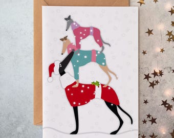 Peut inclure: Une carte de vœux blanche illustrée de trois lévriers empilés portant des pulls. Le chien du bas porte un manteau de Père Noël rouge, celui du milieu un pull bleu sarcelle et celui du haut un pull rose. Des flocons blancs tombent sur le fond gris.