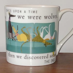 May include: A white ceramic mug featuring a teal sofa illustration with four dogs. The text reads: ONCE UPON A TIME we were wolves... then we discovered sofas THE END. The dogs are depicted in black, yellow, brown, and white colours with collars.