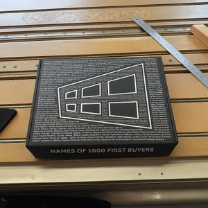 May include: Black box with white geometric design and text. The box has the words "NAMES OF 1000 FIRST BUYERS" printed on the front. A metal ruler is on the wooden surface.