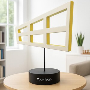 May include: A modern sculpture featuring a white rectangular frame with yellow interior accents, mounted on a black stand. The stand is atop a round wooden table. The sculpture has the text "Your logo" on the base.