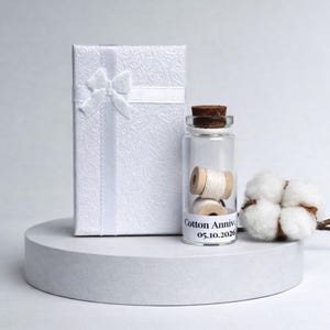 May include: A white gift box with a bow, a small glass bottle with spools of thread, and a cotton branch. The bottle has a cork stopper and a label that reads "Cotton Anniv 05.10.2026". The items are on a white platform.