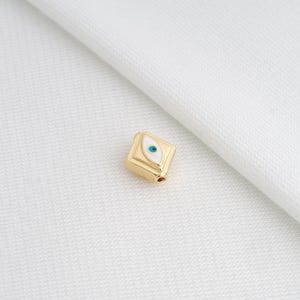 May include: A gold-colored, square bead with an enamel eye design. The eye features a white background with a blue iris. The bead has a small hole for stringing and is set against a white background.