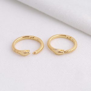 May include: Two gold-colored snake-shaped hoop earrings. The earrings are open, with the snake heads facing each other. The snake's body has a textured scale pattern. The earrings are on a white background.