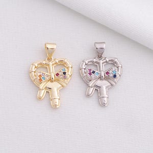 May include: Two heart-shaped pendants with bow accents. One is gold-toned, the other silver-toned. Both feature a key design with a bow adorned with colorful gemstones. The pendants are likely for necklaces or bracelets.