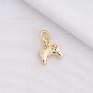 May include: A gold-colored charm in the shape of a video game controller. The charm has a loop at the top for attaching to a bracelet or necklace. The controller has small, colorful gems on the front.