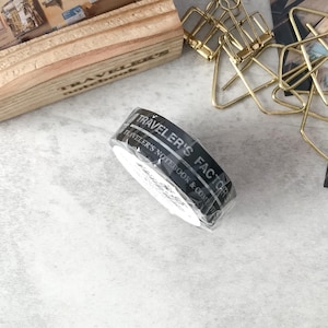 May include: A roll of black washi tape with white text that reads "TRAVELER'S FACTORY" and "TRAVELER'S NOTEBOOK & CO." The tape is partially unwound and sits on a light gray surface. Other stationery items are in the background.