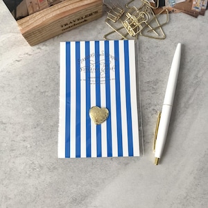 May include: A notebook with blue and white vertical stripes, a gold heart-shaped charm, and a white pen with gold accents. The notebook has text that reads "STANDARD AND STRONG THE SUPERIOR LABOR".