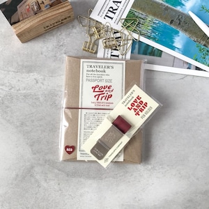 Könnte beinhalten: Ein Reisepass-Notizbuch mit rotem Gummiband, einem roten Stifthalter und einer Holzbox. Das Notizbuch trägt die Aufschrift "Love and Trip". Goldene Büroklammern und Postkarten zum Thema Reisen sind ebenfalls zu sehen.