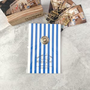 May include: A blue and white striped notebook with a decorative brass button. The notebook has the text "STANDARD AND STRONG THE SUPERIOR LABOR" and "MADE IN JAPAN". Other stationery items are in the background.