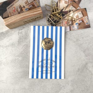 May include: A notebook with blue and white vertical stripes, featuring a brass-colored badge with a bird design. The notebook is labeled "STANDARD AND STRONG THE SUPERIOR LABOR" and "MADE IN JAPAN". Other stationery items are in the background.