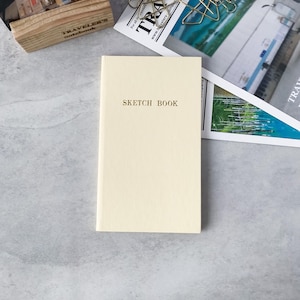 以下が含まれることがあります： クリーム色の表紙に金色の文字でSKETCH BOOKと印刷された長方形のスケッチブック。このノートは、滑らかな質感のシンプルでクリーンなデザインで、平らな面に垂直に配置されています。