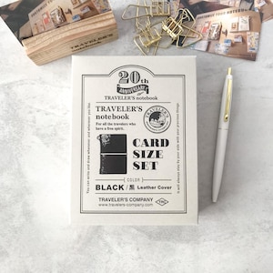 Op de afbeelding: Een zwart-witte kaartset van Traveler's Notebook, met de tekst "20th Anniversary Traveler's Notebook". Een witte pen met gouden accenten ligt naast de set. Andere briefpapierartikelen staan op de achtergrond.