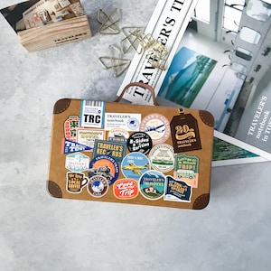 May include: A rectangular brown metal tin case with a handle, decorated with numerous colourful travel-themed stickers featuring text such as Traveler's notebook, Traveler's Airlines, 20th Anniversary, and various illustrations of trains, planes, and coffee.