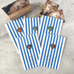 May include: Notebooks with blue and white striped covers are displayed, some with heart-shaped pins that read "HAND MADE." A wooden box with the text "TRAVELER'S NOTEBOOK" is visible, along with gold binder clips and other notebooks.