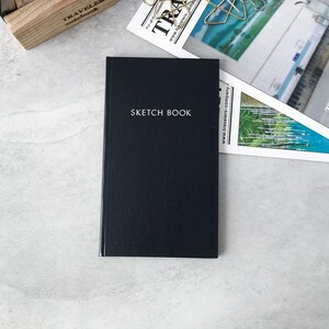 May include: A rectangular black sketchbook with a textured cover and the words SKETCH BOOK printed in white capital letters on the front center. The notebook is positioned vertically, featuring a clean, solid dark surface with a subtle grain.
