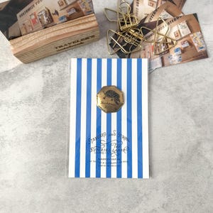 May include: A notebook with blue and white vertical stripes, featuring a gold-colored metal emblem with a turtle design. The notebook has text that reads "STANDARD AND STRONG" and "THE SUPERIOR LABOR". Other items include paper clips and a wooden box.