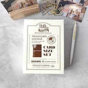 Op de afbeelding: Een wit en bruin Traveler's Notebook kaartformaat set met een bruine lederen cover. De set bevat een notitieboek en is voorzien van de tekst "20th Anniversary Traveler's Notebook". Een witte pen met gouden accenten is ook aanwezig.