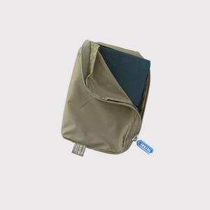 May include: A khaki-colored zippered pouch, partially open to reveal a dark blue object inside. The pouch has a rectangular shape with a zipper closure and a small tag attached. A blue tag with white text is attached to the zipper.