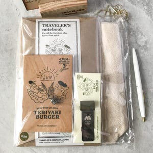 May include: A Traveler's Notebook kit with a brown paper cover, a pen, and a beige fabric roll. The kit includes a pewter charm, a pen holder, and a notebook with the text "TRAVELER'S notebook" and "TERIYAKI BURGER".