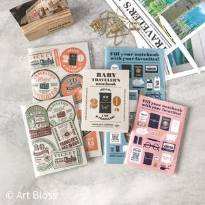 May include: A collection of Traveler's Company notebooks and stationery items featuring commemorative 20th-anniversary designs. The items include various notebooks with illustrated covers, gold-toned metal binder clips, and a wooden display stand.