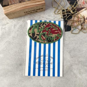Puede incluir: Un parche ovalado con las palabras "Misc. & Crafts" bordadas en verde y rojo. El parche está sobre una tarjeta a rayas azules y blancas con el texto "Standard and Strong". Una caja de madera y clips dorados en el fondo.