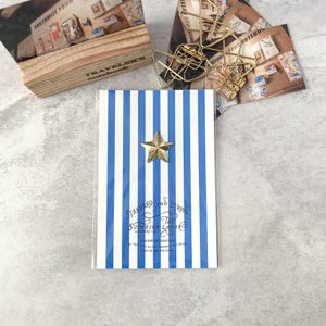 May include: A notebook with blue and white vertical stripes and a gold star emblem. The notebook has the text "STANDARD AND STRONG THE SUPERIOR LABOR" and "MADE IN JAPAN". Other stationery items are in the background.