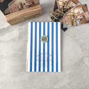 May include: A notebook with a blue and white striped cover. The notebook has a circular pin with the letters "TSL" on it. The text "STANDARD AND STRONG" and "SUPERIOR LABOR" is printed on the cover. The notebook is labeled "MADE IN JAPAN".