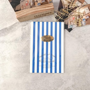 May include: A notebook with blue and white vertical stripes. The notebook has a gold oval label that reads "HELLO T.S.L." The bottom of the notebook has the text "STANDARD AND STRONG THE SUPERIOR LABOR MADE IN JAPAN".