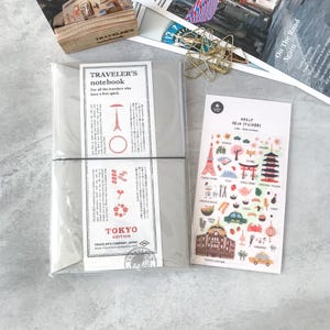 Könnte beinhalten: Ein Traveler's Notizbuch in einer transparenten Plastikhülle mit dem Text "TOKYO EDITION". Daneben ein Bogen "DAILY DECO STICKERS" mit japanischen Illustrationen. Im Hintergrund eine Holzkiste und eine Zeitschrift.