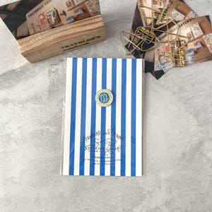 May include: A notebook with blue and white vertical stripes, a gold-colored pin, and text that reads "STANDARD AND STRONG THE SUPERIOR LABOR". A wooden box and gold paper clips are in the background.