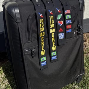 May include: Three black luggage straps with yellow text that reads "10 10 30 IAD CREW" and various airline logos and flags. The straps are attached to a black suitcase.