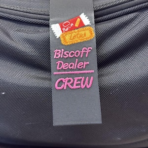 May include: A black bag with a gray strap featuring an embroidered design of a cookie and the words "Biscoff Dealer CREW" in pink. The bag has a zipper and a handle.